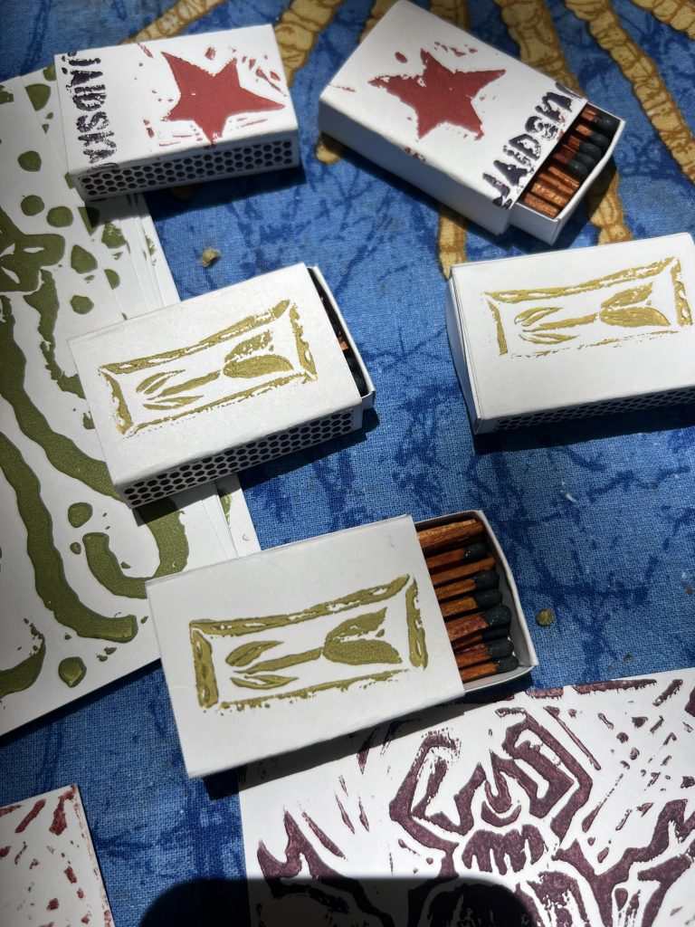 Block printed, handmade matchboxes laying on a table. 3 of the matchboxes have a gold tulip design and 2 have a pink star and "JAIDSKAI" printed on them.